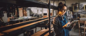 A woman wearing glasses and a denim shirt with a scarf, standing in a workshop and using a tablet. The background features shelves, tools, and wooden materials, indicating a creative or industrial workspace.