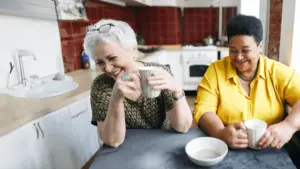 Two friends in a kitchen laugh over a cup of coffee.